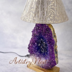 May include: A table lamp with a striking purple amethyst geode base, edged with gold. The lamp has a beige lampshade with a gold pattern and a wooden base. The text "Artistry Nest" is visible.