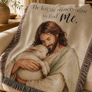 May include: A woven throw blanket featuring a depiction of a bearded figure with long brown hair holding a small white lamb. The text "He left the ninety-nine to find me." is printed in black at the top. The blanket has a fringed edge.
