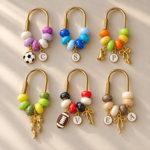 May include: Six gold-toned U-shaped keychains featuring colorful beads and various charms. Charms include a soccer ball, a megaphone, a football, bows, and circular white discs with the letters C, S, P, V, E, and A printed in black.