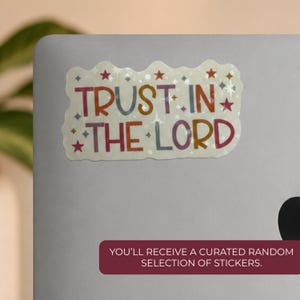 May include: A rectangular sticker with wavy edges featuring the text TRUST IN THE LORD in multi-colored block letters. The background is off-white and decorated with small red, orange, and grey stars and sparkling starburst accents.