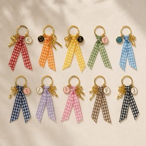 May include: Ten gold-toned metal keychains, each featuring a gingham ribbon in various colors including red, orange, yellow, green, blue, navy, lavender, pink, brown, and black. Each keychain includes a small gold bow charm and a circular letter charm.