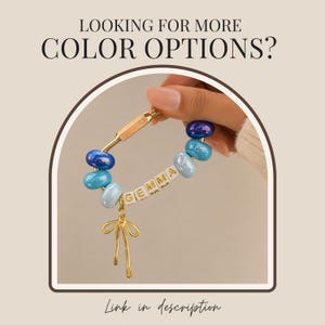May include: A gold-toned carabiner keychain featuring blue and light blue glass beads, white letter beads spelling the name GEMMA, and a gold-toned bow charm. The text LOOKING FOR MORE COLOR OPTIONS? and Link in description are visible.