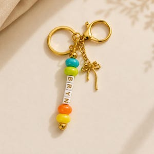 May include: A gold-toned keychain featuring a split ring and a lobster clasp. A vertical strand of beads includes a turquoise bead, a lime green bead, four white square beads spelling BRYN, an orange bead, and a yellow bead. A gold bow charm hangs nearby.