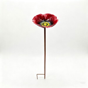 May include: A red poppy garden stake with a black center and yellow accents. The flower is made of a shiny material and is mounted on a brown metal pole with a ground stake.