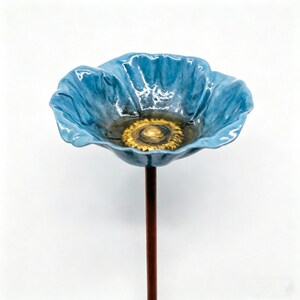 May include: A ceramic blue poppy flower with a golden center, mounted on a brown metal stem. The flower has a glossy finish and detailed petal edges. The poppy is a decorative garden accent.