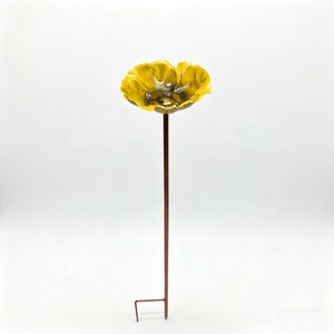 May include: A yellow flower-shaped garden stake with a brown stem and a small, square foot. The flower has a textured surface and a dark center. The stake is designed to be placed in the ground.