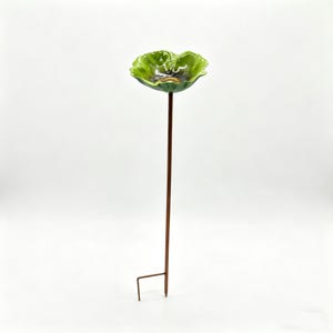 May include: A green ceramic bird bath with a flower-shaped bowl and a brown metal stake. The bowl has a glossy finish and a dark center. The stake has a ground anchor for easy placement in a garden or yard.