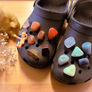 May include: A pair of dark gray clogs adorned with various colorful gemstones and a small plastic figurine. The gemstones include orange, red, green, and blue hues. A small, seated Buddha statue and dried flowers are also present.