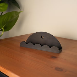 May include: A dark grey, semi-circular wall-mounted organiser with a scalloped bottom edge. The organiser has a small hole at the top and is placed on a wooden surface. A green plant is visible in the background.