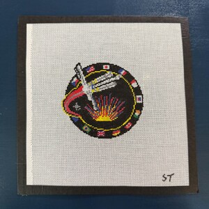 May include: Needlepoint art piece featuring a black circle with a hand and flame design, surrounded by small flags. The artwork is on white canvas, framed in a dark brown border. The initials "ST" are in the lower right corner.