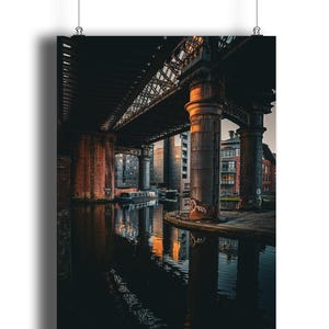 May include: A vertical print of a canal scene under a bridge. The image features a dark, moody aesthetic with reflections in the water. The bridge's pillars and the water's surface reflect the warm light of the setting sun.