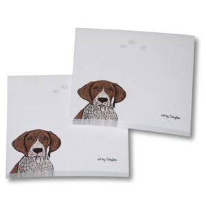 May include: Two white square notepads with a brown and white dog illustration. The dog's head is centered on the notepad, with paw prints in the upper corner. The text "art by Sabylaa" is in the lower right corner.