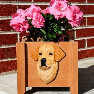 May include: A square wooden planter box featuring a carved, three-dimensional golden retriever head on the front panel. The planter is filled with a vibrant plant displaying numerous bright pink flowers with green leaves and stems.