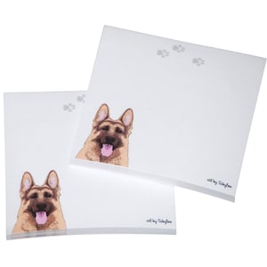 May include: Two packs of notepad paper with a German Shepherd dog illustration on the front. The dog is brown and black with a pink tongue sticking out. The text "art by Sabyloo" is printed on the bottom right corner of each notepad.