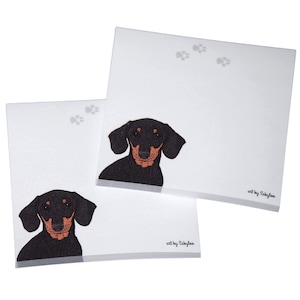 May include: Two white notepads with a black and tan dachshund illustration on each. The notepads have a gray paw print design and the text "art by Sakylos" at the bottom.