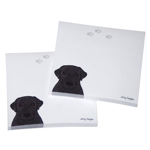 May include: Two white notepads with black paw prints and a black Labrador Retriever illustration. The notepad on the bottom has the text "art by Schuyler" at the bottom right corner. The notepad on top has the text "art by Schuyler" at the bottom right corner.