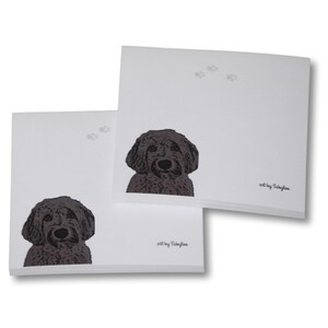 May include: Two white square notepads with a dark brown dog illustration and paw prints. The dog's head is centered on the bottom of the notepad. The artist's signature, "art by Sabylaa", is in the bottom right corner.