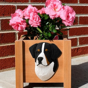 Peut inclure: Une jardinière carrée en bois brun clair ornée d'un relief en trois dimensions représentant une tête de bouvier bernois sur le panneau avant. La jardinière est remplie d'un bouquet vibrant de fleurs d'azalées roses et de feuilles vertes.