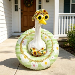 May include: A white goose statue wearing a sunflower-patterned bucket hat, yellow flower-shaped sunglasses, and a matching sunflower-patterned bikini top, positioned inside a light green and white striped inflatable pool float with white flowers.