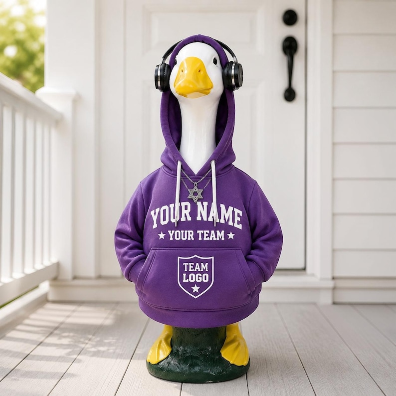 May include: A white goose statue wearing a purple hooded sweatshirt with white text that reads YOUR NAME, YOUR TEAM, and TEAM LOGO inside a shield graphic. The goose wears black over-ear headphones and a silver Star of David pendant necklace.