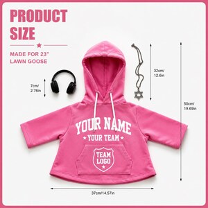 May include: A bright pink hooded sweatshirt with white drawstrings, featuring the text 'YOUR NAME', 'YOUR TEAM', and 'TEAM LOGO' in white. Includes black headphones and a silver Star of David necklace. Dimensions are 37cm/14.57in wide and 50cm/19.69in tall.