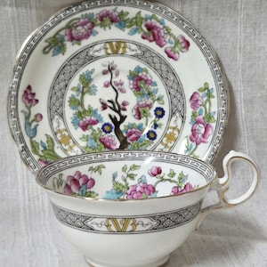 May include: A white teacup and saucer set with a floral design in pink, blue, and green. The set features gold trim and a decorative border. The saucer is approximately 15 cm in diameter.