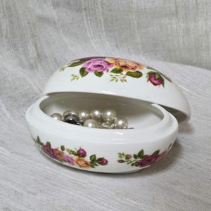 May include: Oval white ceramic jewelry box decorated with pink, yellow, and green roses. The open box reveals pearls and a silver ring. The lid is slightly ajar, showcasing the floral pattern on the inside.