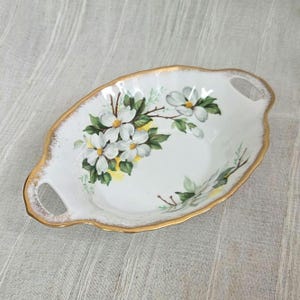 May include: Oval white ceramic serving dish with gold trim and handles. The dish features a floral pattern of white flowers, green leaves, and yellow accents. A decorative piece for serving, approx. 25 cm long.
