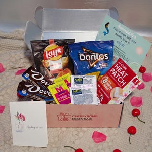 Peut inclure: Une boîte cadeau en carton marquée Cherrybomb Essentials contenant des chips barbecue Lay's, des Doritos Cool Ranch, des barres de chocolat noir Dove, des sachets d'Advil et un patch chauffant JJ Care. Une carte Mermaid Era et une carte Thinking of You.