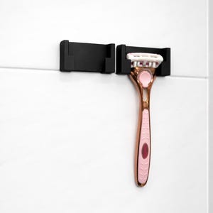 May include: A black razor holder with a rose gold and pink razor. The razor has a pink handle with a rose gold accent and a white blade head. The holder is mounted on a white tiled wall.
