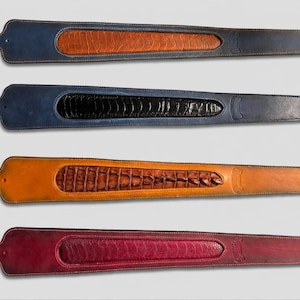 May include: Four leather guitar straps arranged vertically. Each strap features a wide, tapered body with a textured, inlaid center panel. The straps are colored navy blue with tan, navy blue with black, tan with brown, and deep red.