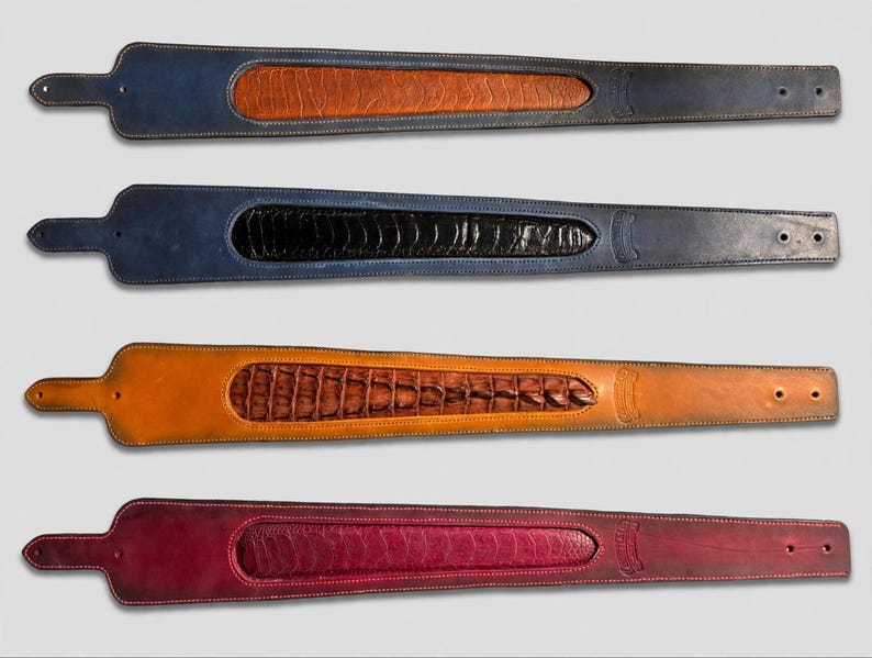 May include: Four leather guitar straps arranged vertically. Each strap features a wide, tapered body with a textured, inlaid center panel. The straps are colored navy blue with tan, navy blue with black, tan with brown, and deep red.