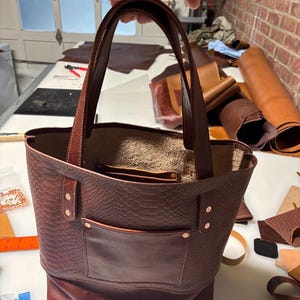 May include: A dark brown leather tote bag featuring a textured snakeskin pattern on the upper portion and a smooth leather exterior pocket. The bag includes two sturdy leather handles, copper-toned rivets, and an open top design.