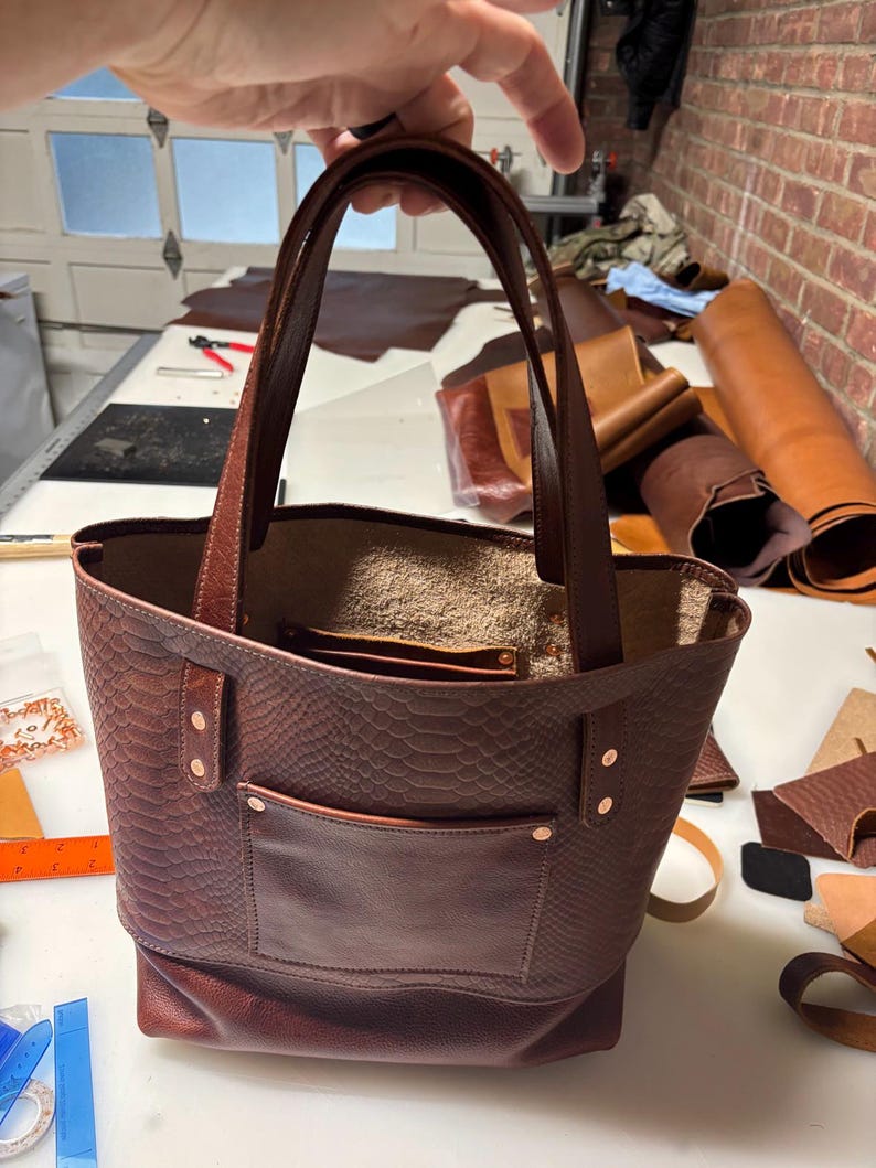 May include: A dark brown leather tote bag featuring a textured snakeskin pattern on the upper portion and a smooth leather exterior pocket. The bag includes two sturdy leather handles, copper-toned rivets, and an open top design.