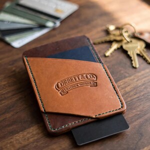 May include: A tan and dark brown leather card holder featuring green stitching and the embossed text Corbett & Co. Leather Works. The wallet has a slanted front pocket and holds a black card, with additional cards and keys nearby.