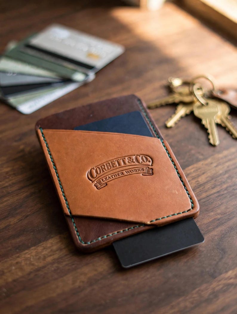 May include: A tan and dark brown leather card holder featuring green stitching and the embossed text Corbett & Co. Leather Works. The wallet has a slanted front pocket and holds a black card, with additional cards and keys nearby.