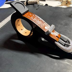 May include: A tan leather belt with a repeating embossed pattern, featuring a silver-toned metal buckle and a matching silver-toned metal keeper. The belt is coiled and mounted on a black display stand, with a ruler visible in the background.