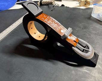 May include: A tan leather belt with a repeating embossed pattern, featuring a silver-toned metal buckle and a matching silver-toned metal keeper. The belt is coiled and mounted on a black display stand, with a ruler visible in the background.