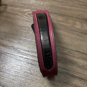 May include: A belt featuring a central black panel with a textured, reptilian-like pattern, bordered by smooth, deep magenta leather edges. The design includes visible stitching along the length of the magenta trim, creating a structured appearance.