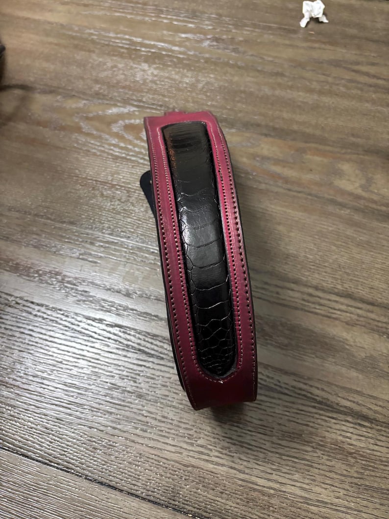 May include: A belt featuring a central black panel with a textured, reptilian-like pattern, bordered by smooth, deep magenta leather edges. The design includes visible stitching along the length of the magenta trim, creating a structured appearance.