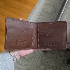 May include: A brown leather bifold wallet featuring multiple card slots on both sides. The left side is embossed with the text Corbett & Co. Leather Works, while the right side is embossed with the text Semper Fidelis. The wallet has visible stitching.