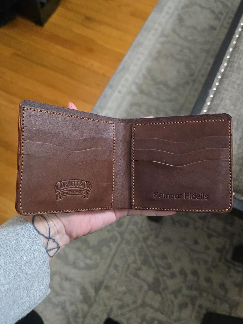 May include: A brown leather bifold wallet featuring multiple card slots on both sides. The left side is embossed with the text Corbett & Co. Leather Works, while the right side is embossed with the text Semper Fidelis. The wallet has visible stitching.