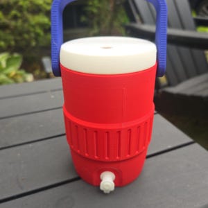 May include: A red and white water cooler with a blue handle. The cooler has a white spigot and a white lid. The handle is arched and the cooler is sitting on a dark gray surface.