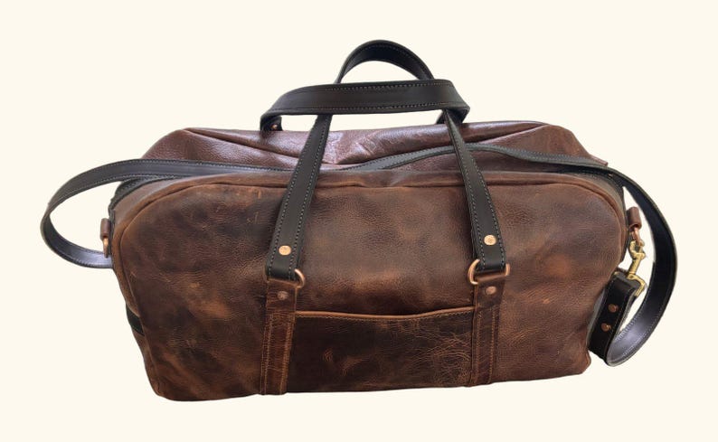 Op de afbeelding: Een bruine leren reistas met donkerbruine leren handvatten en een bijpassende schouderriem. De tas is voorzien van een voorvak, messingkleurige metalen hardware en zichtbare stiksels bij de bevestigingspunten van de handvatten.