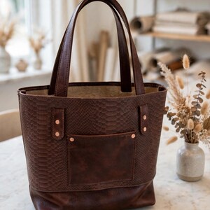 May include: A dark brown tote bag featuring a snakeskin-textured upper body and a smooth leather lower section. The bag includes two tall handles, a front pocket with copper-colored rivets, and a spacious open top with a light-colored interior.