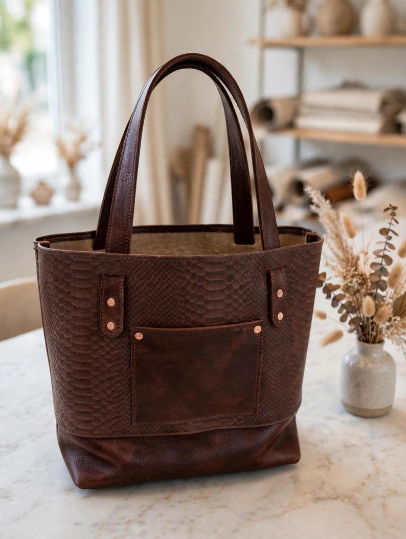 May include: A dark brown tote bag featuring a snakeskin-textured upper body and a smooth leather lower section. The bag includes two tall handles, a front pocket with copper-colored rivets, and a spacious open top with a light-colored interior.