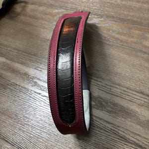 May include: A curved belt featuring a central black panel with a textured, reptilian-like pattern, bordered by smooth, deep magenta leather edges. The belt displays visible stitching along the length of the magenta trim and has a light-colored interior lining.