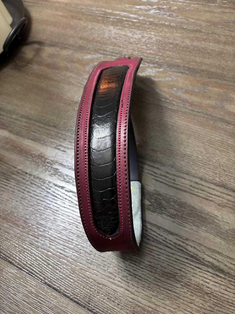 May include: A curved belt featuring a central black panel with a textured, reptilian-like pattern, bordered by smooth, deep magenta leather edges. The belt displays visible stitching along the length of the magenta trim and has a light-colored interior lining.