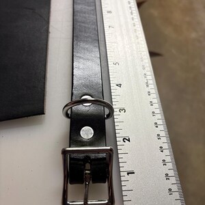 May include: A black leather strap with a silver-toned metal buckle and a D-ring, positioned next to a metal ruler showing measurements in inches and centimeters to indicate the length of the strap and hardware components.