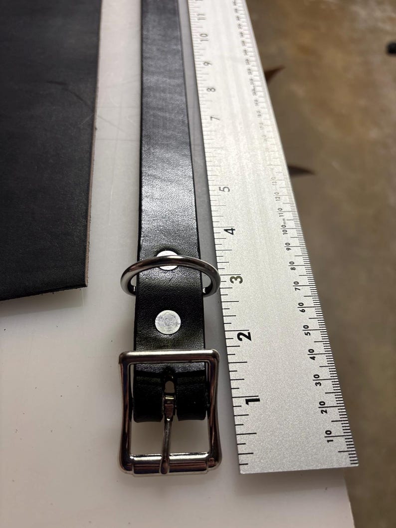 May include: A black leather strap with a silver-toned metal buckle and a D-ring, positioned next to a metal ruler showing measurements in inches and centimeters to indicate the length of the strap and hardware components.